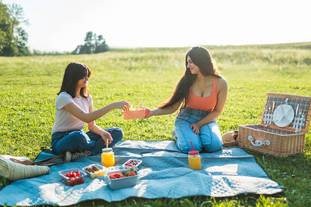 Young diverse female friends are sitting on a blanket in a sunny meadow, enjoying a picnic with snacks and drinks. The scene is featuring a serene outdoor setting with a picnic basket and vibrant greenery.