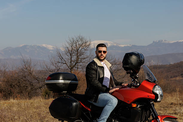 Handsome young biker in leather jacket sitting on his motorcycle while taking a break from ride in sunny nature