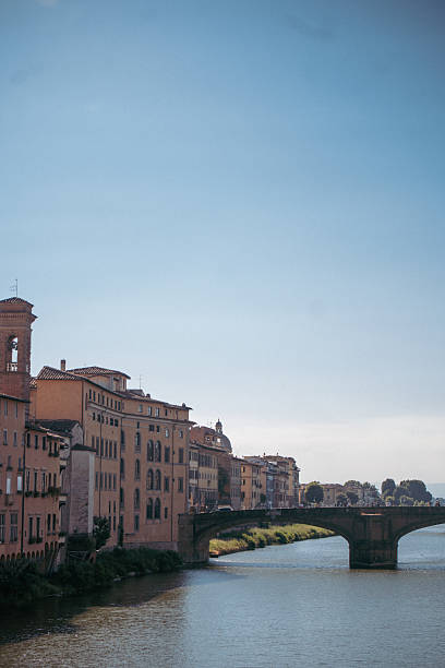 Wide view along the Arno River with historic buildings, bridges, and gentle river traffic under a bright sky, capturing classic urban scenery and relaxed atmosphere in Florence, Italy.