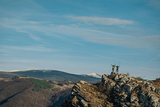 Two hikers celebrating their achievement at the mountain summit, taking in a breathtaking scenic view of valleys and forests below