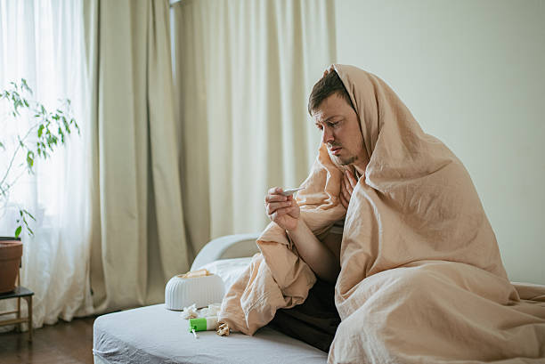 istock Man resting in bed, covered with blanket, measuring temperature with thermometer, surrounded by tissues and medication, recuperating during illness 2232465737