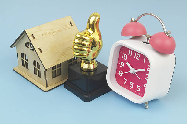 Alarm clock, house model and thumbs up on blue background.