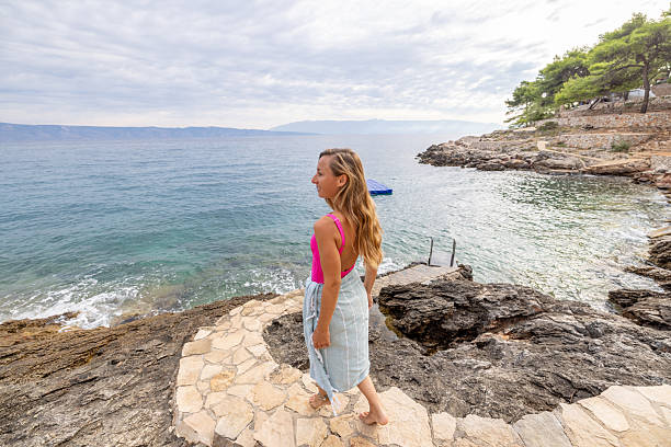 Woman in pink top and skirt walking on stone path by rocky shore toward calm sea. Scenic coastal landscape with clear water and distant hills. Outdoor nature and travel concept.