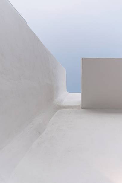 Minimalist view of a whitewashed building in Chora Donousa, Cyclades, Greece, captured from a looking-up perspective. The abstract lines and soft geometry highlight traditional Cycladic architecture against the clear blue summer sky. Perfect for themes of travel, Mediterranean lifestyle, architecture, and minimalism.