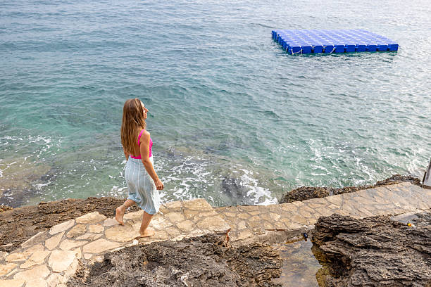 Woman in pink top and skirt walking on stone path by rocky shore toward calm sea. Scenic coastal landscape with clear water and distant hills. Outdoor nature and travel concept.