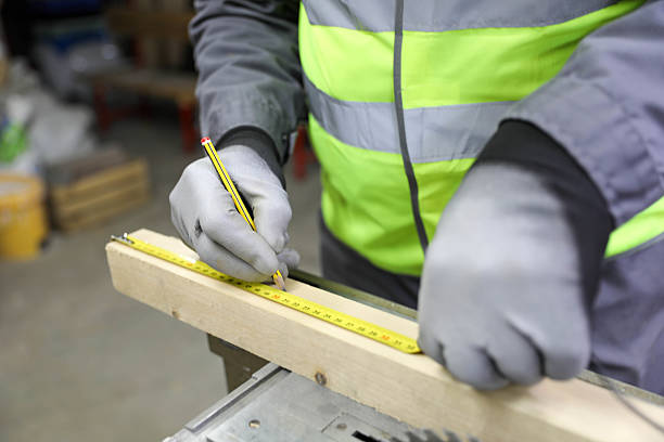 Construction Worker in Protective Gloves and Reflective Safety Vest Measuring Wooden Plank with Tape Measure Near Circular Saw in Workshop. Concept of Carpentry, Precision, and Skilled Labor in Industrial Environment.