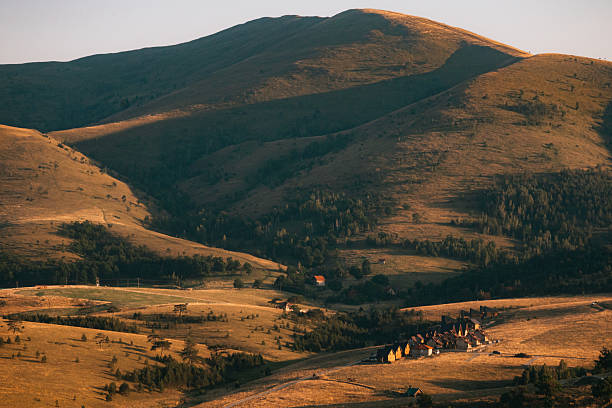 Scenic view of a small village surrounded by rolling golden hills and dramatic mountain landscape at sunset.