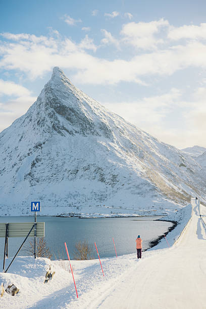 Tourist in distance wearing pink jacket exploring Lofoten in Northern Norway during wintertime, admiring scenic snowy landscape of bridge and the beautiful mountain