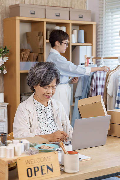 Senior asian mother smiling while working on laptop preparing shop orders as adult asian daughter arranging clothing rack during family fashion boutique small business ecommerce teamwork management