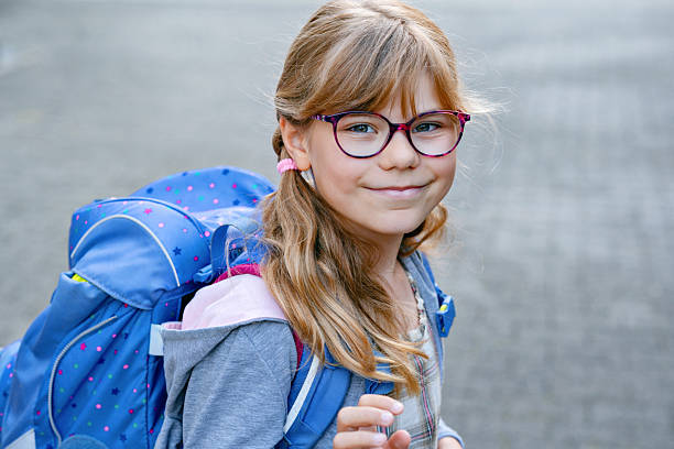istock Happy little elementary school girl with eyeglasses and satchel backpack walking outdoors on the way to school. Smiling pupil child ready for back to school, education and childhood lifestyle concept. 2232492877