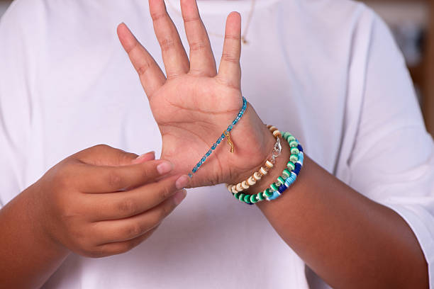 Close-up of a child’s hands adjusting a delicate blue beaded bracelet with a small golden charm while wearing other colorful handmade bracelets on the wrist. The plain white shirt background highlights the jewelry, creativity, and do-it-yourself accessory crafting process.