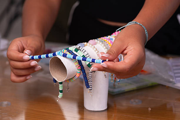 Close-up of hands holding a blue and white handmade beaded bracelet above a display stand filled with multiple colorful bracelets. The scene includes jewelry-making materials in the background, representing creativity, handmade accessories, and do-it-yourself fashion design.