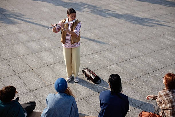 Teenage Caucasian girl standing outdoors playing flute for small group of seated people, musical instrument case lying on ground nearby, audience watching performance attentively