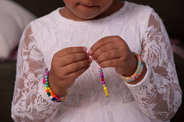 Close-up of a young girl’s hands carefully threading bright multicolored beads onto a string while wearing colorful bracelets and a white lace dress. The image highlights focus, patience, and the joy of childhood creativity through jewelry-making in a home environment.