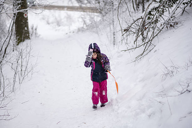 Young girl pulling an orange sled through the snowy woods, dressed in warm winter clothes, enjoying a fun outdoor adventure in nature