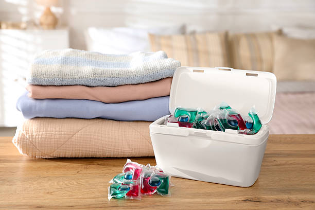 istock Detergent capsules in container and clean laundry on wooden table indoors 2232498102