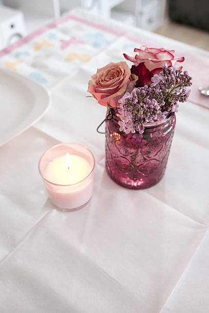 Close-up of a decorative table arrangement featuring a pink glass jar with roses and lilac flowers alongside a lit candle. The soft light and pastel colors create a cozy and romantic atmosphere.