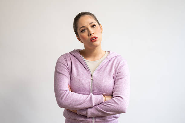 A surprised young Caucasian female is standing with her arms crossed, wearing a casual pink hoodie. She is looking directly at the camera with a neutral background, featuring a simple and minimalist setting.