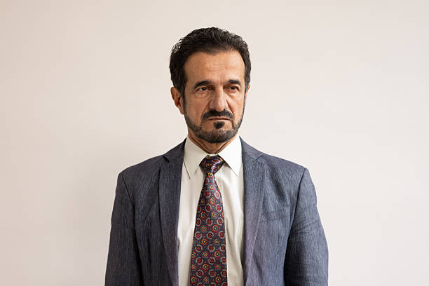 A serious senior Middle Eastern businessman in a formal suit and patterned tie is standing against a plain background. He is exuding confidence and professionalism in a corporate setting.