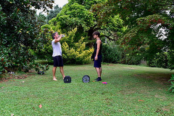 Two individuals engage in a fitness session in a lush garden, one demonstrating strength training with weights while being filmed by the other.