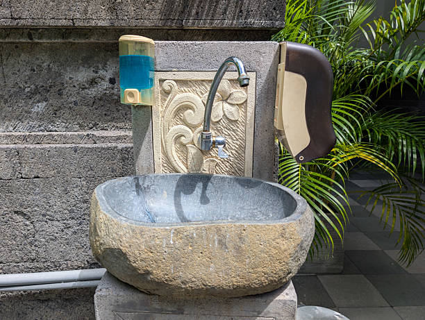 A natural stone bowl-shaped sink equipped with a faucet, liquid soap dispenser, and tissue holder, mounted on a wall with traditional ornamental carvings.