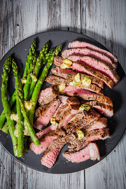 Sliced grilled steak served alongside steamed asparagus, garnished with butter and herbs. The dish is presented on a black ceramic plate placed over a rustic wooden surface. Perfectly cooked, this flavorful meal displays a balance of textures and colors, evoking a sense of gourmet dining and culinary delight.