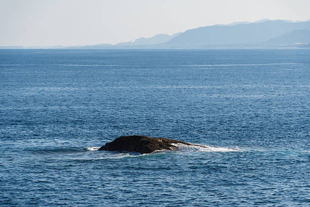 A solitary rock emerging from the calm blue sea with distant mountains on the horizon, creating a peaceful coastal landscape.