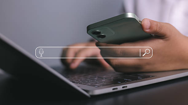 istock Person holds a smartphone while typing on a laptop keyboard, with a floating search bar interface, symbolizing modern search engine optimization, digital marketing, and the use of internet technology. 2232537654