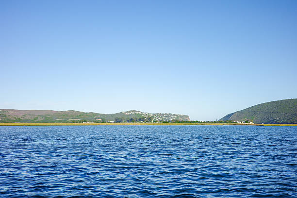 Calm blue ocean with rolling green hills and a distant coastal community under a bright clear sky. A peaceful and scenic seascape, travel, nature, lifestyle, relaxation, and coastal in Knysna
