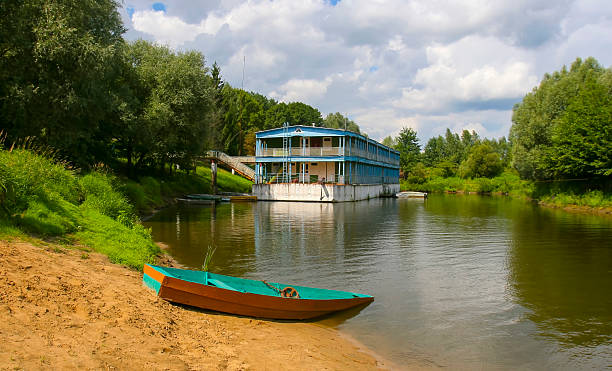 istock Boat on sandy shore near blue building reflecting in calm river under cloudy sky 2232548292