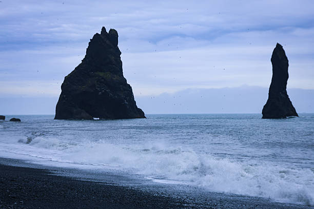 Waves crash against the volcanic black sand shore with Reynisdrangar sea stacks rising from the Atlantic Ocean near Vík í Mýrdal, Iceland. This iconic coastal scene captures the raw power and beauty of Iceland’s southern coastline, where geology, mythology, and dramatic landscapes meet. A stunning travel and nature image symbolizing adventure, exploration, and natural wonder.