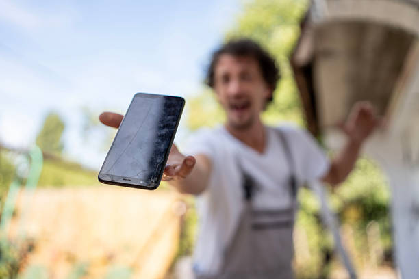 A person focuses attention on a cracked smartphone in focus, held out in a foreground setting against a blurred outdoor background, portraying damage and interest in smartphone repair.