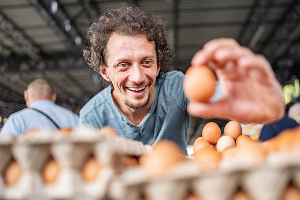 A joyful man carefully choosing eggs at a bustling farmers market, showcasing fresh and organic produce.