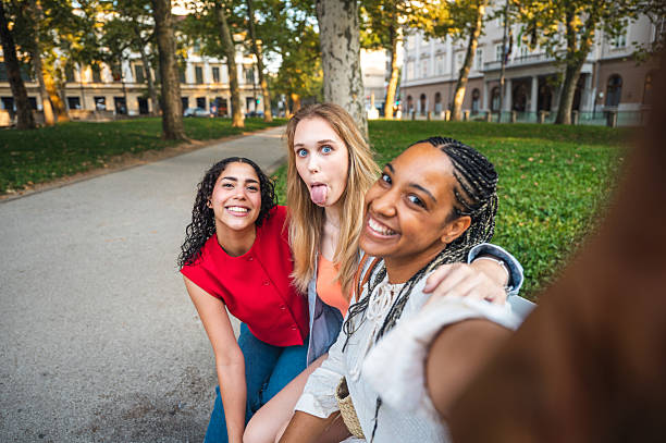 Young diverse female friends are sitting on a park bench having fun taking a selfie, featuring a vibrant urban park setting with lush greenery and historic buildings in the background.