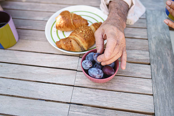 A senior Hispanic male is sitting at a wooden table, enjoying pastries and fresh plums. His hand is reaching for a bowl of ripe plums, featuring a relaxed outdoor setting with a plate of croissants.
