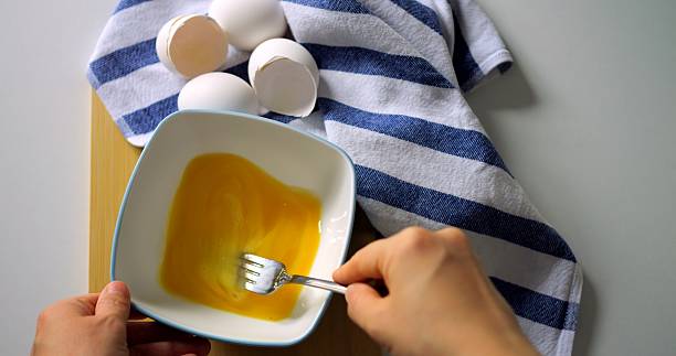 istock Closeup of hand whisking egg mixture in white bowl with fork, striped kitchen towel and egg shells nearby, top view breakfast preparation. 2232575338