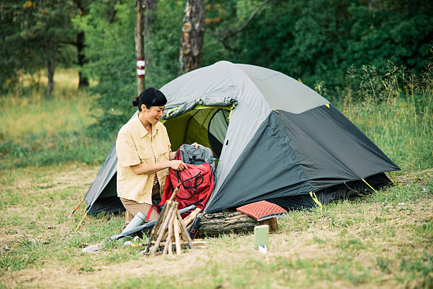 A camper organizes her gear near a modern tent amidst a peaceful forest. Essential camping items are visible. The scene highlights outdoor leisure and preparation for nature excursions.