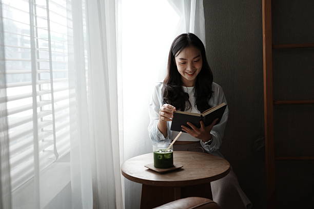 Young Asian woman enjoying a matcha latte and reading a book by a window in a cozy cafe, embracing a peaceful moment of relaxation
