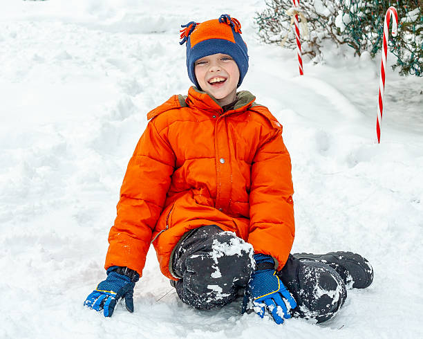 Front view of smiling cute boy sitting outdoors in the snow. Holiday decorations in background.