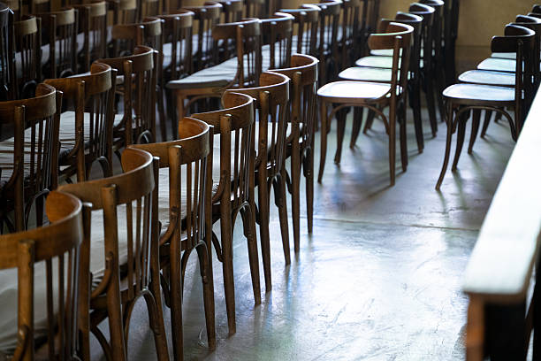 Neatly arranged rows of classic wooden chairs stand in an empty hall, with light reflecting softly on the polished floor. The curved backs create a repeating pattern, adding rhythm and perspective to the scene. The image conveys anticipation, order, and the quiet atmosphere before an event or gathering.