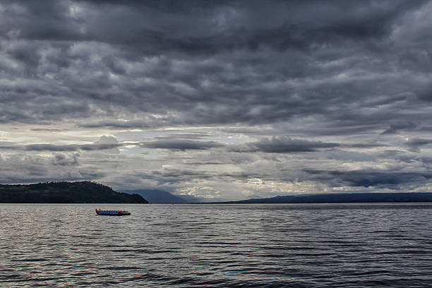 A scenic view of a calm lake under a dramatic cloudy sky, with mountains and hills visible in the background. The moody atmosphere creates a sense of tranquility mixed with intensity, symbolizing nature’s beauty, weather changes, and peaceful solitude. A floating inflatable raft adds a small focal point to the wide natural landscape. Suitable for themes of travel, adventure, nature scenery, weather, outdoor activities, and landscape photography