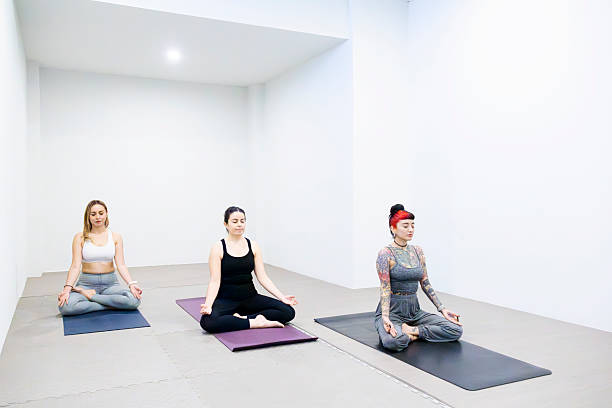 A group of multi-ethnic adults sit with their legs crossed and their arms raised above their heads with hands in a prayer pose. They each have neutral expressions on their faces.  The focus is on a Hispanic woman with her hair pulled back and comfortable active wear on.