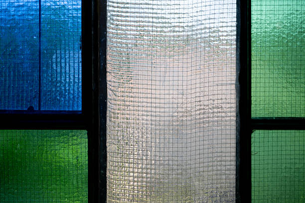 Close-up of a stained glass window with textured panels in blue, green, and clear tones. The grid-like surface diffuses the light, creating a soft and abstract effect. The image emphasizes geometry, color, and the artistic detail of architectural design.