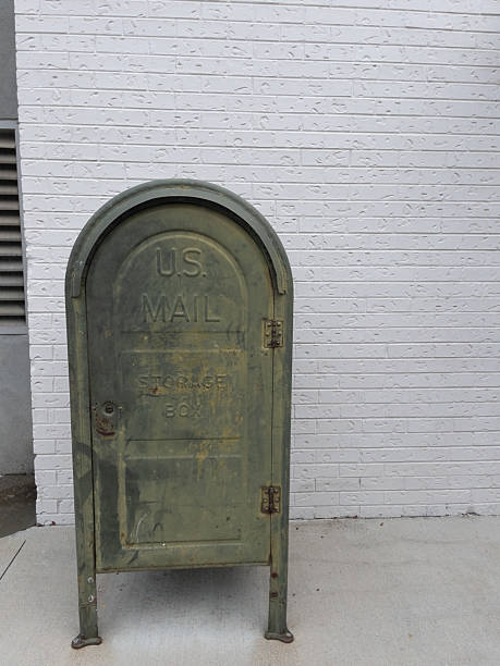 Old, weathered, green U.S. Mail storage box stands on legs outside against a white brick wall