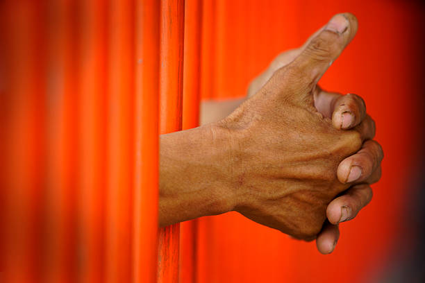 Close-up of hands gripping orange bars, symbolizing confinement and struggle. The image captures emotion and the complex relationship between hope and isolation.