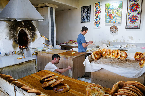 Tashkent, Uzbekistan August 04 2025: A local bread vendor at Chorsu Bazaar in Tashkent, Uzbekistan, proudly displays freshly baked round loaves of traditional Uzbek bread. Captured in the heart of the bustling market, the artisan is seen both crafting and selling the bread, embodying the rich culinary heritage of Central Asia. The warm tones and authentic setting highlight the cultural significance of bread in Uzbek daily life.