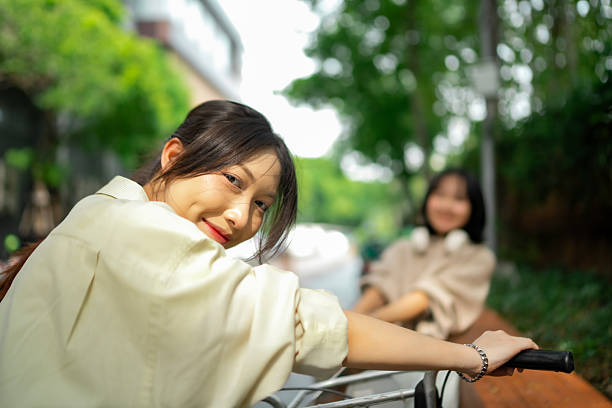 Two girls enjoy time together near a busy road, one confidently guiding her bicycle while the other shows playful happiness. Their smiles reflect energy, friendship, confidence, and urban exploration.