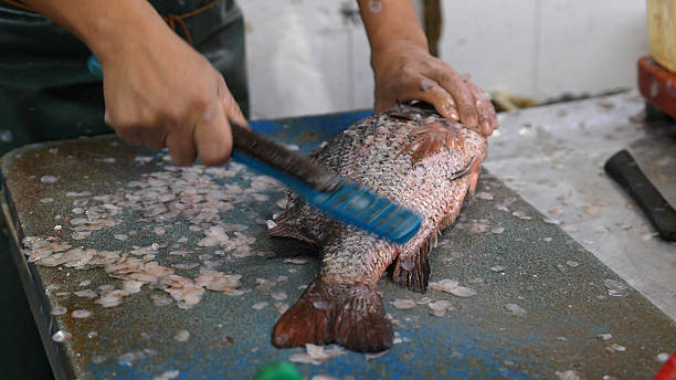 Close-up of a vendor descaling fresh fish on a chopping board at a seafood market, showing hands and preparation process.