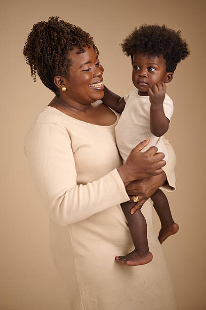 A warm portrait of a smiling mother holding her young child against a neutral beige background. The woman, dressed in a light cream outfit, embraces her toddler, who wears a simple beige onesie.