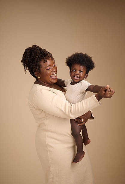 A candid portrait of a mother playfully holding her toddler against a neutral beige background. The woman, dressed in a light cream outfit, looks joyfully at her child, who is pointing outward.